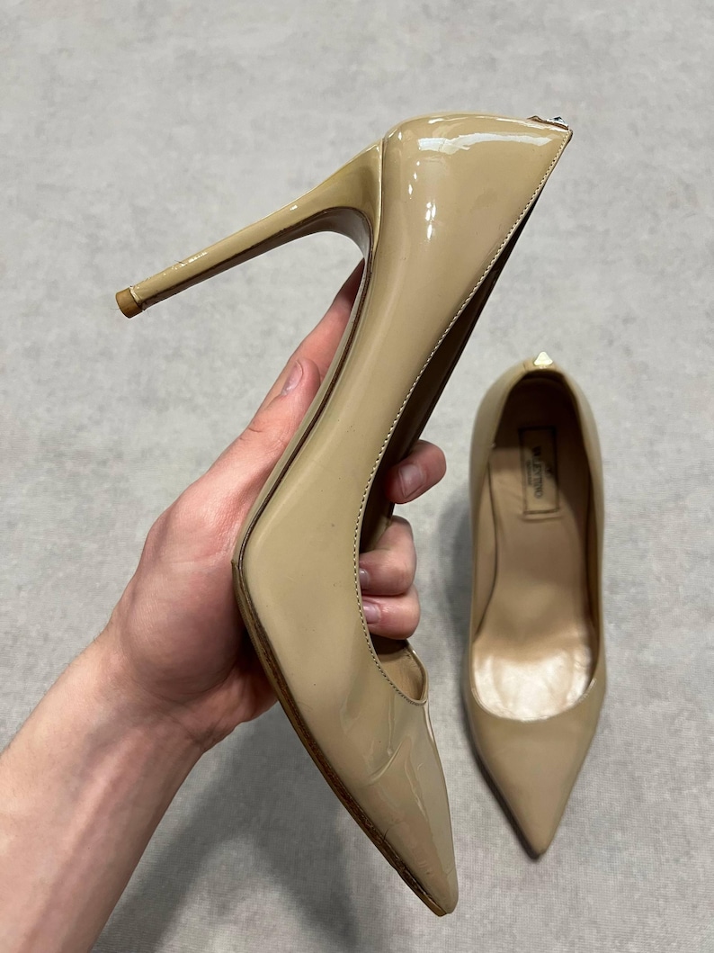 Puede incluir: Zapatos de tac&oacute;n alto de charol beige. Se sostiene un zapato, mostrando el tac&oacute;n de aguja y la punta afilada. El otro zapato est&aacute; en el fondo. Los zapatos tienen un acabado brillante y un peque&ntilde;o detalle dorado.