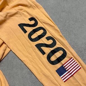 May include: Mustard-colored long-sleeved shirt with the number "2020" printed in large black letters on the sleeve. An American flag is printed below the numbers. The shirt is made of a soft material.