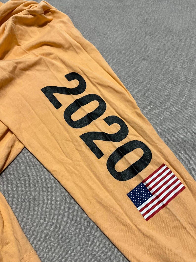 May include: Mustard-colored long-sleeved shirt with the number "2020" printed in large black letters on the sleeve. An American flag is printed below the numbers. The shirt is made of a soft material.