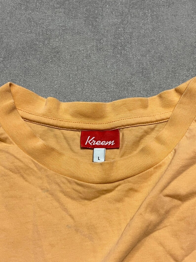 May include: A light orange t-shirt with a crew neck. The red tag at the neck reads "Kreem" in white lettering. The shirt is a size large.