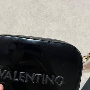 May include: A glossy black clutch with the embossed word "VALENTINO" visible. The clutch has a rectangular shape with rounded edges and a gold-colored chain strap. The background is a neutral, light gray color.
