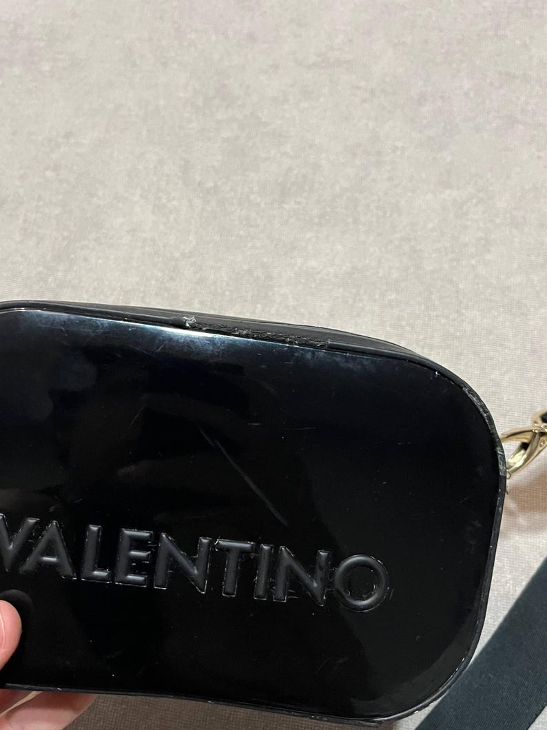 May include: A glossy black clutch with the embossed word "VALENTINO" visible. The clutch has a rectangular shape with rounded edges and a gold-colored chain strap. The background is a neutral, light gray color.