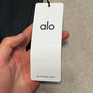 May include: A rectangular white paper hang tag attached by a black string to a dark garment. The tag features the black lowercase text alo centered at the top and the website address ALOYOGA.COM printed in black at the bottom of the tag.