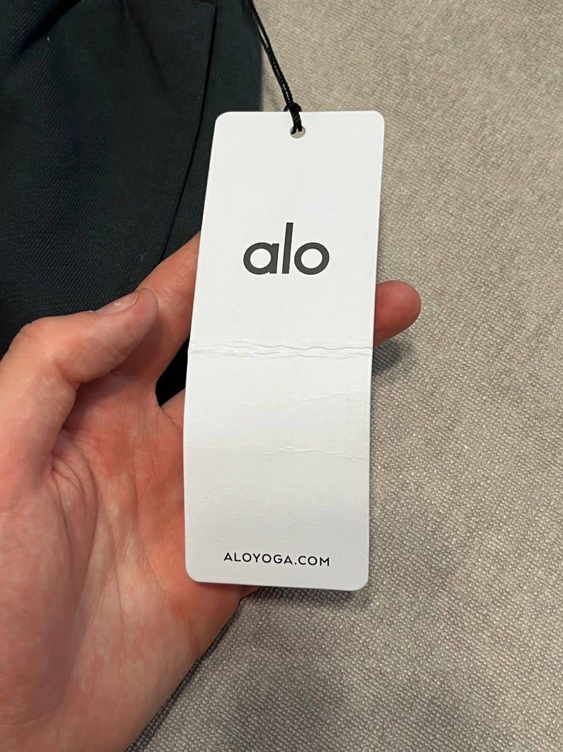 May include: A rectangular white paper hang tag attached by a black string to a dark garment. The tag features the black lowercase text alo centered at the top and the website address ALOYOGA.COM printed in black at the bottom of the tag.
