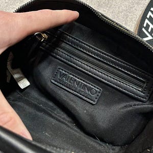May include: Black crossbody bag with a zippered main compartment and an interior zippered pocket. The bag features a gold-toned zipper and the "VALENTINO MARIO VALENTINO SPA" logo on a black leather patch.