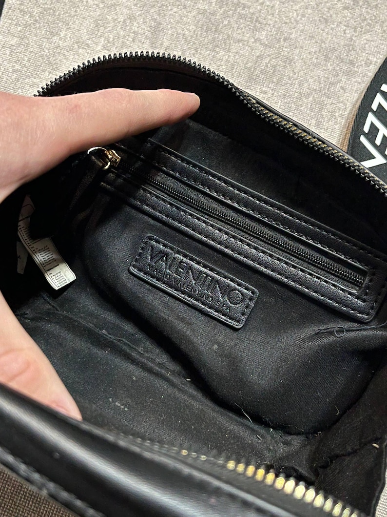May include: Black crossbody bag with a zippered main compartment and an interior zippered pocket. The bag features a gold-toned zipper and the "VALENTINO MARIO VALENTINO SPA" logo on a black leather patch.