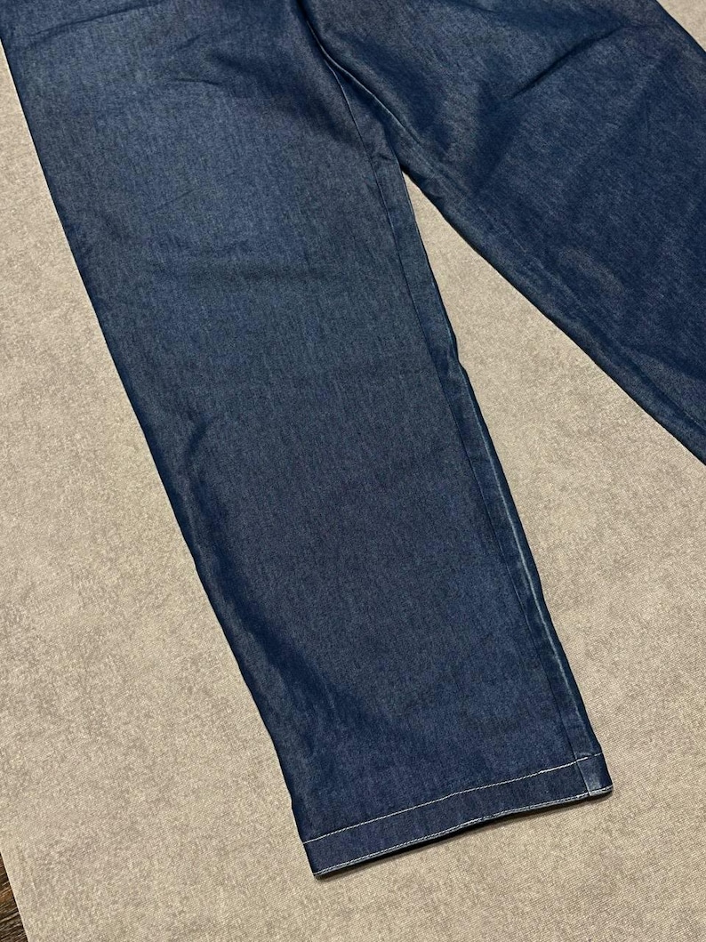 Puede incluir: Primer plano de unos vaqueros de mezclilla azul medio. Los vaqueros tienen un corte de pierna recta y est&aacute;n hechos de una tela de mezclilla resistente. El dobladillo est&aacute; acabado con una costura blanca.