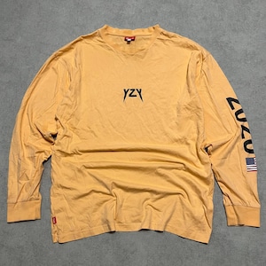May include: Mustard yellow long-sleeve shirt with the text "YZY" in black on the chest. The left sleeve has the text "CUZ" in black and a small American flag. The shirt has a crew neck and a small red tag near the hem.