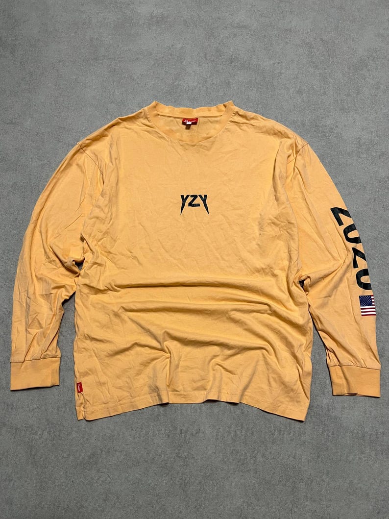 May include: Mustard yellow long-sleeve shirt with the text "YZY" in black on the chest. The left sleeve has the text "CUZ" in black and a small American flag. The shirt has a crew neck and a small red tag near the hem.