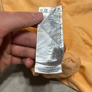 May include: Close-up of a peach-colored garment tag with text in multiple languages. The tag is held by a hand. The tag includes washing instructions and fabric composition details.