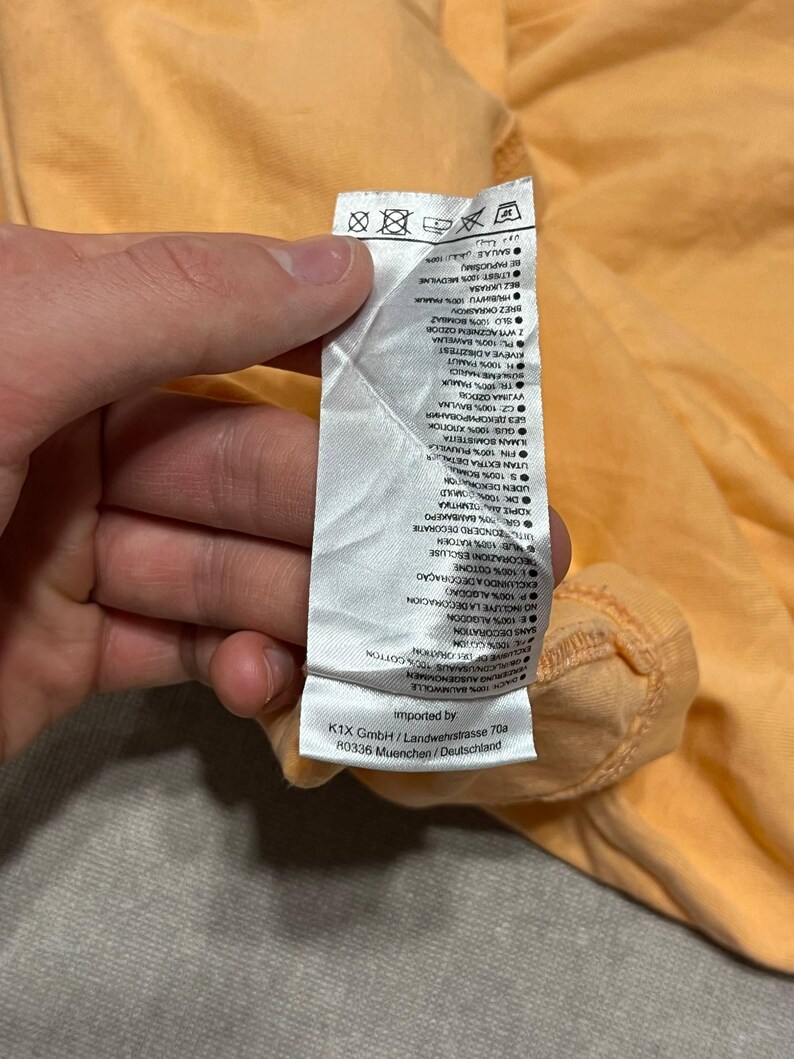 May include: Close-up of a peach-colored garment tag with text in multiple languages. The tag is held by a hand. The tag includes washing instructions and fabric composition details.