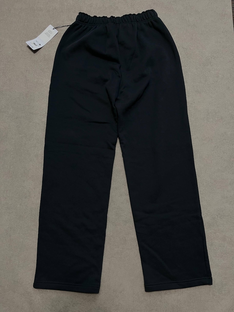 May include: Solid black trousers featuring a high-rise elasticated waistband with a gathered texture. The pants have a straight-leg cut and a clean, unadorned finish. A white rectangular retail tag is attached to the waistband by a black string.