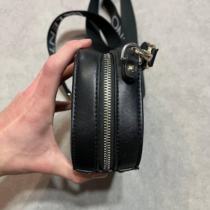 May include: Black, round crossbody bag with a silver zipper and a black strap. The strap has white text that reads "ONLY". The bag is held by a hand.