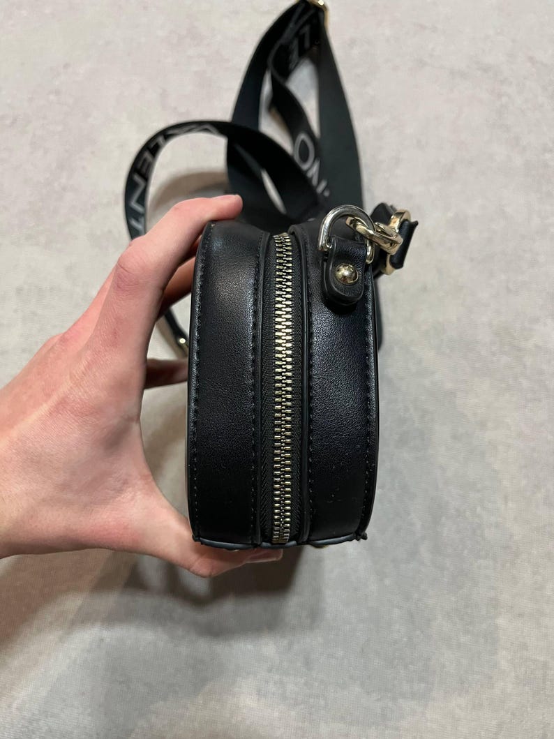 May include: Black, round crossbody bag with a silver zipper and a black strap. The strap has white text that reads "ONLY". The bag is held by a hand.