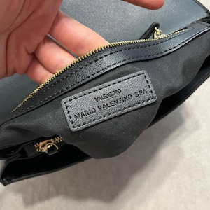 May include: Black leather handbag with a gold zipper and the brand name "VALENTINO MARIO VALENTINO SPA" embossed on a black leather tag. The interior is lined with black fabric.