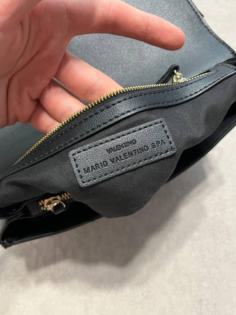 May include: Black leather handbag with a gold zipper and the brand name "VALENTINO MARIO VALENTINO SPA" embossed on a black leather tag. The interior is lined with black fabric.