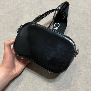 May include: A black, glossy, rectangular crossbody bag with a wide strap. The strap is black with white text. The bag has gold-colored hardware. The bag is held by a hand.