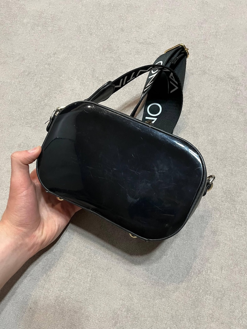 May include: A black, glossy, rectangular crossbody bag with a wide strap. The strap is black with white text. The bag has gold-colored hardware. The bag is held by a hand.