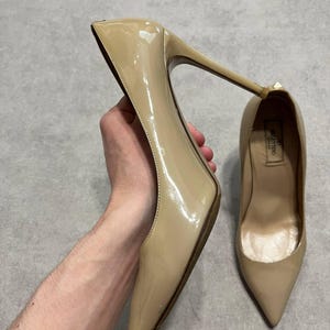 Puede incluir: Un par de zapatos de tac&oacute;n alto de charol beige. Un zapato se sostiene, mostrando el tac&oacute;n de aguja y la punta afilada. El otro zapato est&aacute; sobre una superficie plana. La marca "Valentino" es visible dentro del zapato.