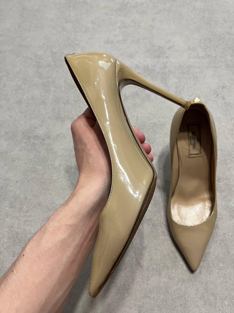 Puede incluir: Un par de zapatos de tac&oacute;n alto de charol beige. Un zapato se sostiene, mostrando el tac&oacute;n de aguja y la punta afilada. El otro zapato est&aacute; sobre una superficie plana. La marca "Valentino" es visible dentro del zapato.