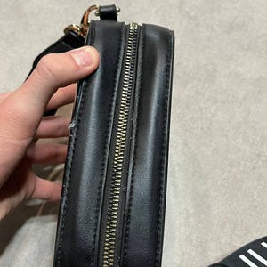 May include: Black rectangular crossbody bag with a gold zipper and strap hardware. The bag has a black strap with white text. The bag is made of a smooth, possibly leather-like material.