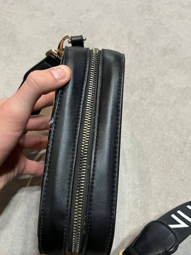 May include: Black rectangular crossbody bag with a gold zipper and strap hardware. The bag has a black strap with white text. The bag is made of a smooth, possibly leather-like material.