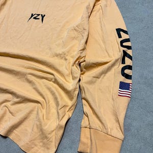 May include: A long-sleeved, peach-colored shirt with the word "YZY" in black script on the chest. The right sleeve features the number "2020" in black and a small American flag. The shirt has a ribbed cuff and a crew neck.