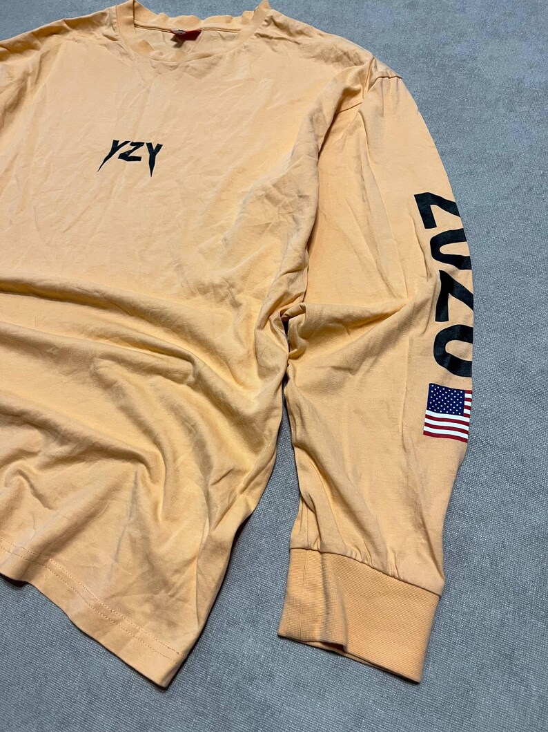 May include: A long-sleeved, peach-colored shirt with the word "YZY" in black script on the chest. The right sleeve features the number "2020" in black and a small American flag. The shirt has a ribbed cuff and a crew neck.