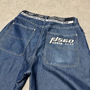 Puede incluir: Vaqueros azules con bolsillos traseros grandes y una cinturilla blanca con el texto "JEANS URBAN GEAR 560". El bolsillo trasero tiene un logotipo blanco que dice "FJ 560 URBAN GEAR".