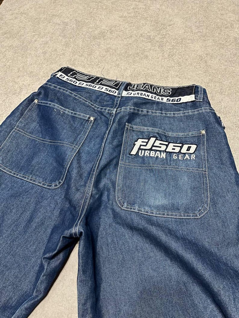 Puede incluir: Vaqueros azules con bolsillos traseros grandes y una cinturilla blanca con el texto "JEANS URBAN GEAR 560". El bolsillo trasero tiene un logotipo blanco que dice "FJ 560 URBAN GEAR".