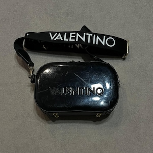 May include: Black Valentino crossbody bag with a matching strap. The bag has a rectangular shape with rounded edges and a zipper closure. The word "VALENTINO" is printed in white on the strap and the bag.
