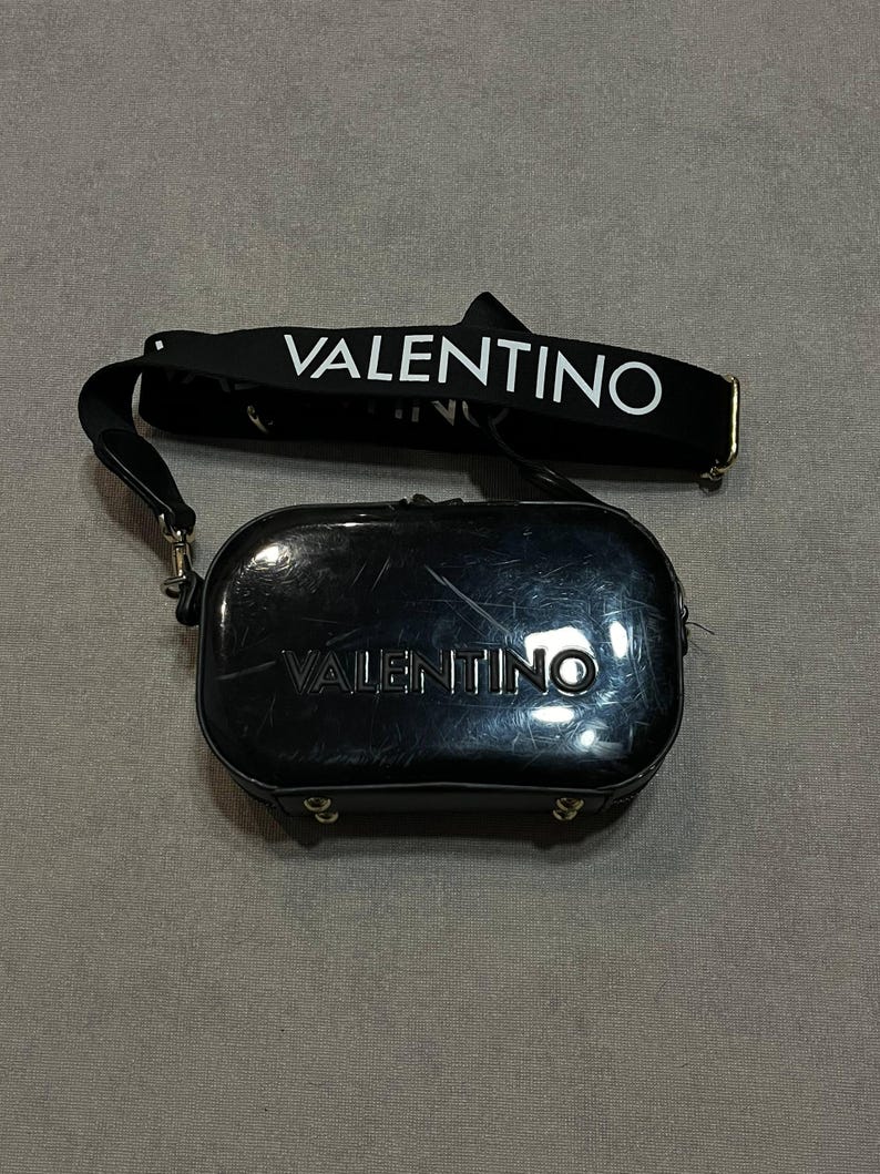 May include: Black Valentino crossbody bag with a matching strap. The bag has a rectangular shape with rounded edges and a zipper closure. The word "VALENTINO" is printed in white on the strap and the bag.