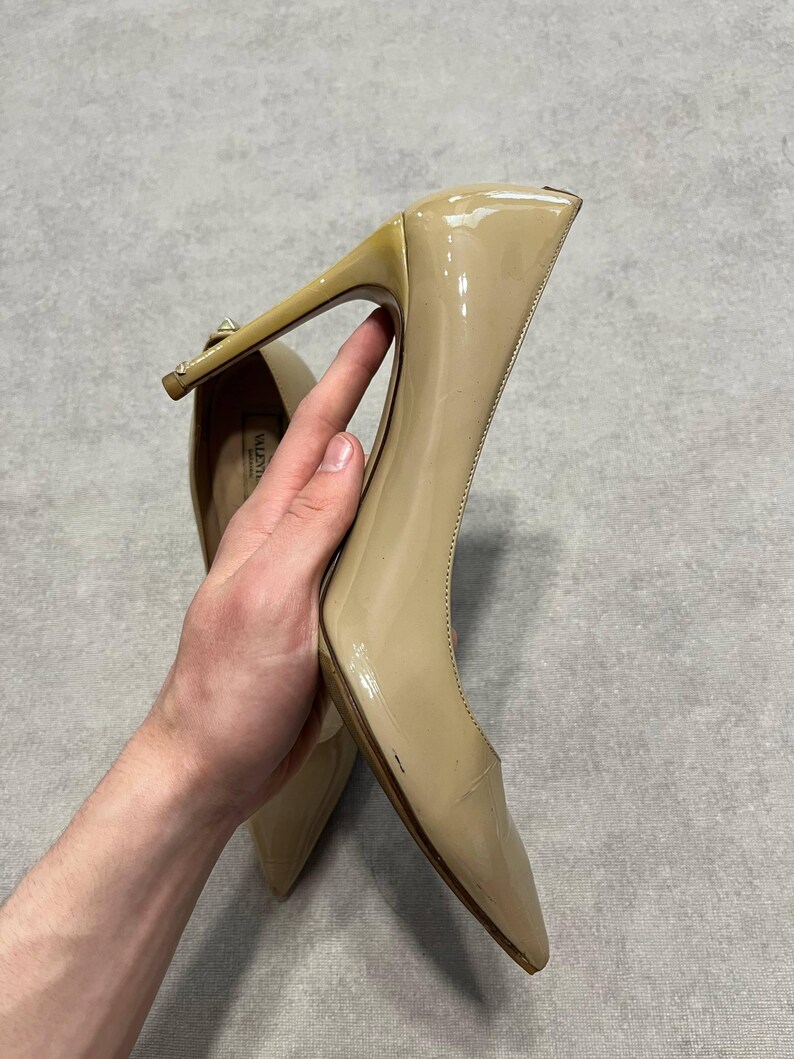 Puede incluir: Un par de zapatos de tac&oacute;n de charol beige. Los zapatos tienen punta afilada y tac&oacute;n de aguja. Los tacones miden aproximadamente 10 cm de alto. Los zapatos son sostenidos por una persona.