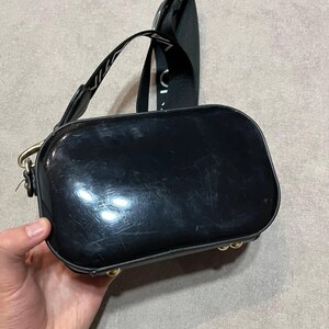 May include: A glossy black crossbody bag with a rectangular shape and rounded edges. The bag has a black strap with white text and gold-colored hardware. The bag is held by a hand.