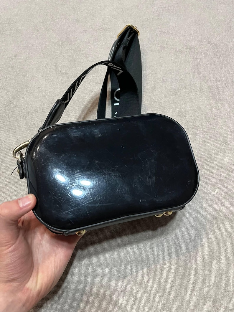 May include: A glossy black crossbody bag with a rectangular shape and rounded edges. The bag has a black strap with white text and gold-colored hardware. The bag is held by a hand.