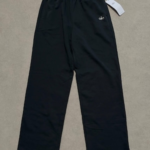 May include: Black sweatpants featuring a high-waisted elastic waistband, side pockets, and a straight-leg silhouette. The word alo is embroidered in white on the upper left thigh. A white brand tag is attached to the waistband with a black string.