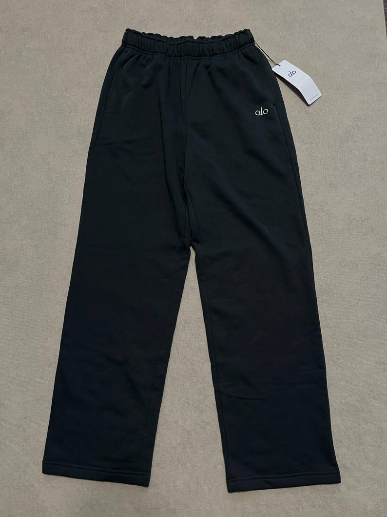 May include: Black sweatpants featuring a high-waisted elastic waistband, side pockets, and a straight-leg silhouette. The word alo is embroidered in white on the upper left thigh. A white brand tag is attached to the waistband with a black string.