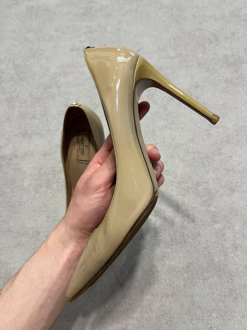 Puede incluir: Un par de zapatos de tac&oacute;n de charol beige. Los zapatos de tac&oacute;n tienen una punta afilada y un tac&oacute;n de aguja. El nombre de la marca "Valentino" es visible en la plantilla de un zapato. Los zapatos son sostenidos por una persona.