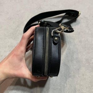 May include: Black crossbody bag with a silver zipper and gold-colored hardware. The bag has a black strap and is made of a smooth material. The bag is held by a hand.