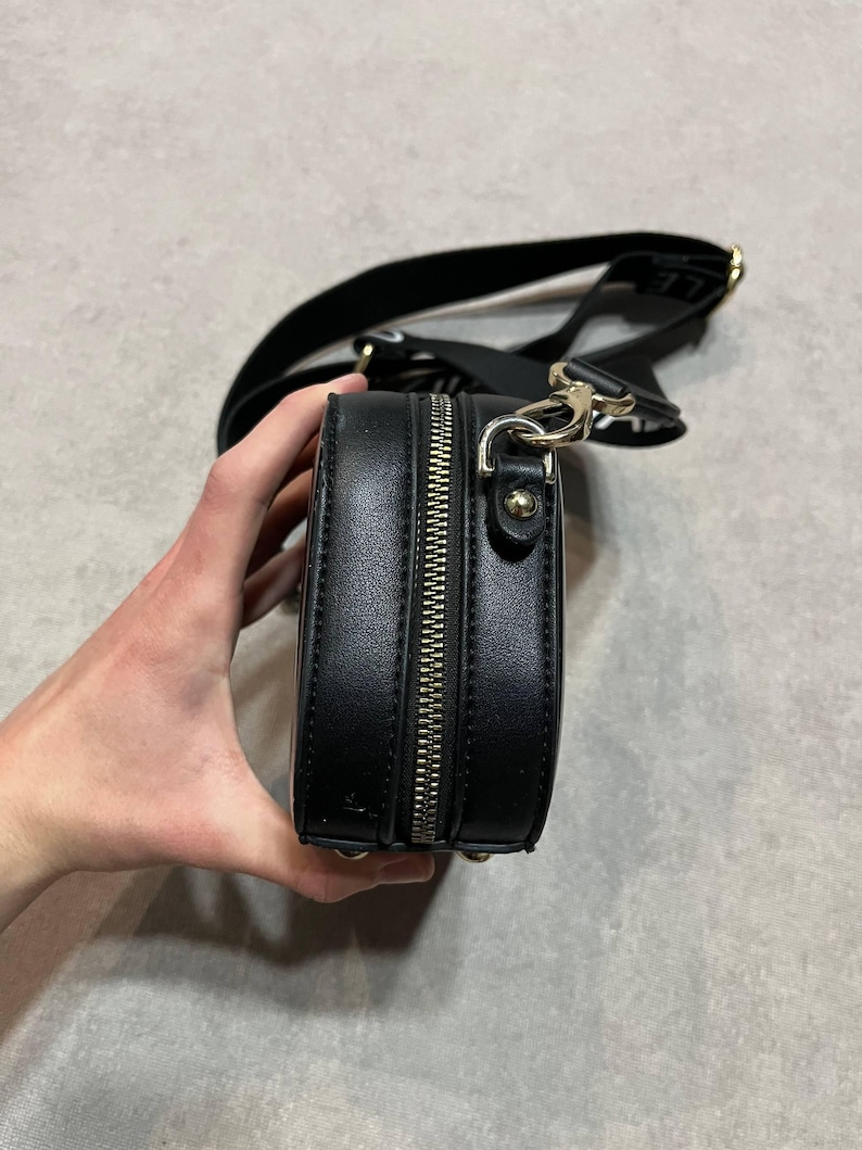 May include: Black crossbody bag with a silver zipper and gold-colored hardware. The bag has a black strap and is made of a smooth material. The bag is held by a hand.