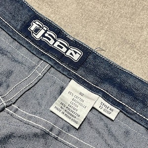 Puede incluir: Primer plano de unos vaqueros de mezclilla azules con costuras blancas. Los vaqueros tienen una etiqueta que dice "TJ560" y una etiqueta de talla que indica la talla 32. La tela es una mezclilla de lavado medio con una textura visible. El n&uacute;mero de estilo es FJ-0018P.