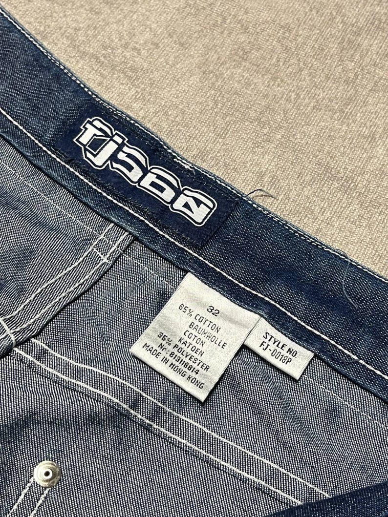 Puede incluir: Primer plano de unos vaqueros de mezclilla azules con costuras blancas. Los vaqueros tienen una etiqueta que dice "TJ560" y una etiqueta de talla que indica la talla 32. La tela es una mezclilla de lavado medio con una textura visible. El n&uacute;mero de estilo es FJ-0018P.