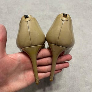 Puede incluir: Un par de tacones altos de charol beige. Los zapatos tienen punta y tac&oacute;n de aguja. Cada tac&oacute;n presenta un peque&ntilde;o acento met&aacute;lico oscuro en la parte superior. Los zapatos est&aacute;n sostenidos por una mano.