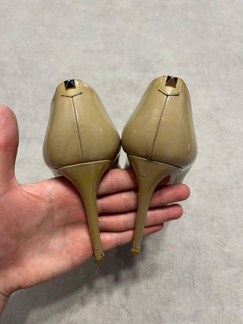 Puede incluir: Un par de tacones altos de charol beige. Los zapatos tienen punta y tac&oacute;n de aguja. Cada tac&oacute;n presenta un peque&ntilde;o acento met&aacute;lico oscuro en la parte superior. Los zapatos est&aacute;n sostenidos por una mano.