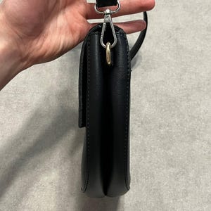May include: A black crossbody bag with a flap closure. The bag has a silver-tone metal clasp and a black strap. The bag is held by a hand against a neutral background.