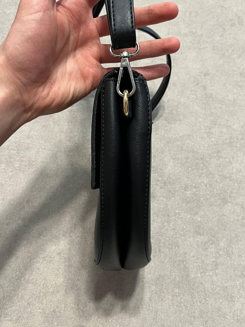 May include: A black crossbody bag with a flap closure. The bag has a silver-tone metal clasp and a black strap. The bag is held by a hand against a neutral background.