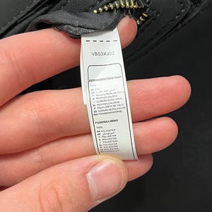May include: Close-up of a black handbag with a gold zipper. A white tag with the text "VBS3XJ02" and material composition details is visible. The bag's exterior is made of PVC, and the lining is polyester.