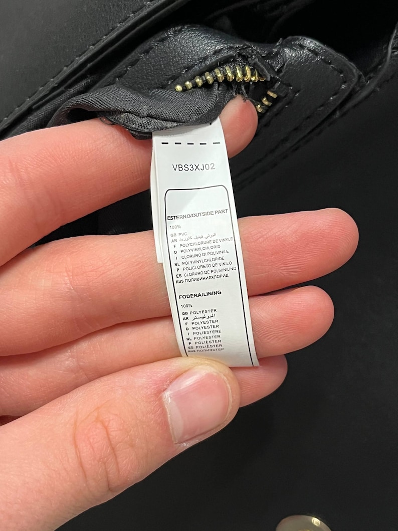 May include: Close-up of a black handbag with a gold zipper. A white tag with the text "VBS3XJ02" and material composition details is visible. The bag's exterior is made of PVC, and the lining is polyester.