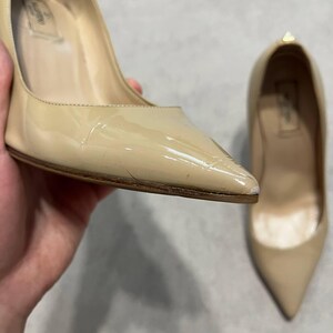 Puede incluir: Zapatos de tac&oacute;n de charol beige con punta. Un zapato se sostiene en la mano, el otro est&aacute; cerca. Los zapatos tienen un tac&oacute;n alto y un acabado brillante. El nombre de la marca es visible en la plantilla.