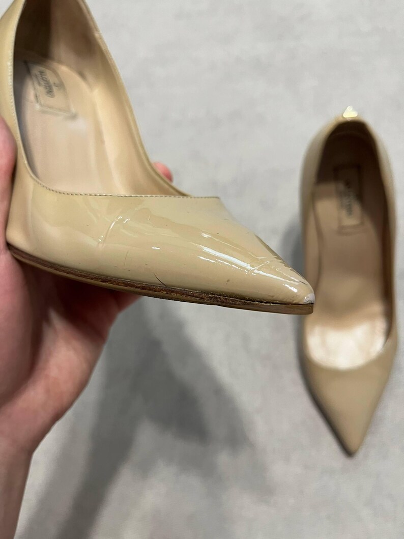 Puede incluir: Zapatos de tac&oacute;n de charol beige con punta. Un zapato se sostiene en la mano, el otro est&aacute; cerca. Los zapatos tienen un tac&oacute;n alto y un acabado brillante. El nombre de la marca es visible en la plantilla.